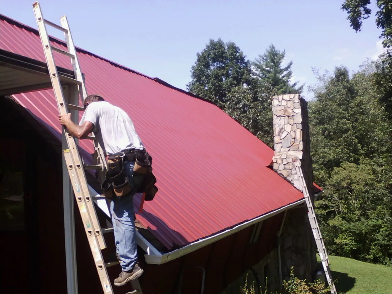 Roofer on a ladder beside a red metal roof and stone chimney — Soffit & Fascia Repair by Roofing Brothers in Cimarron